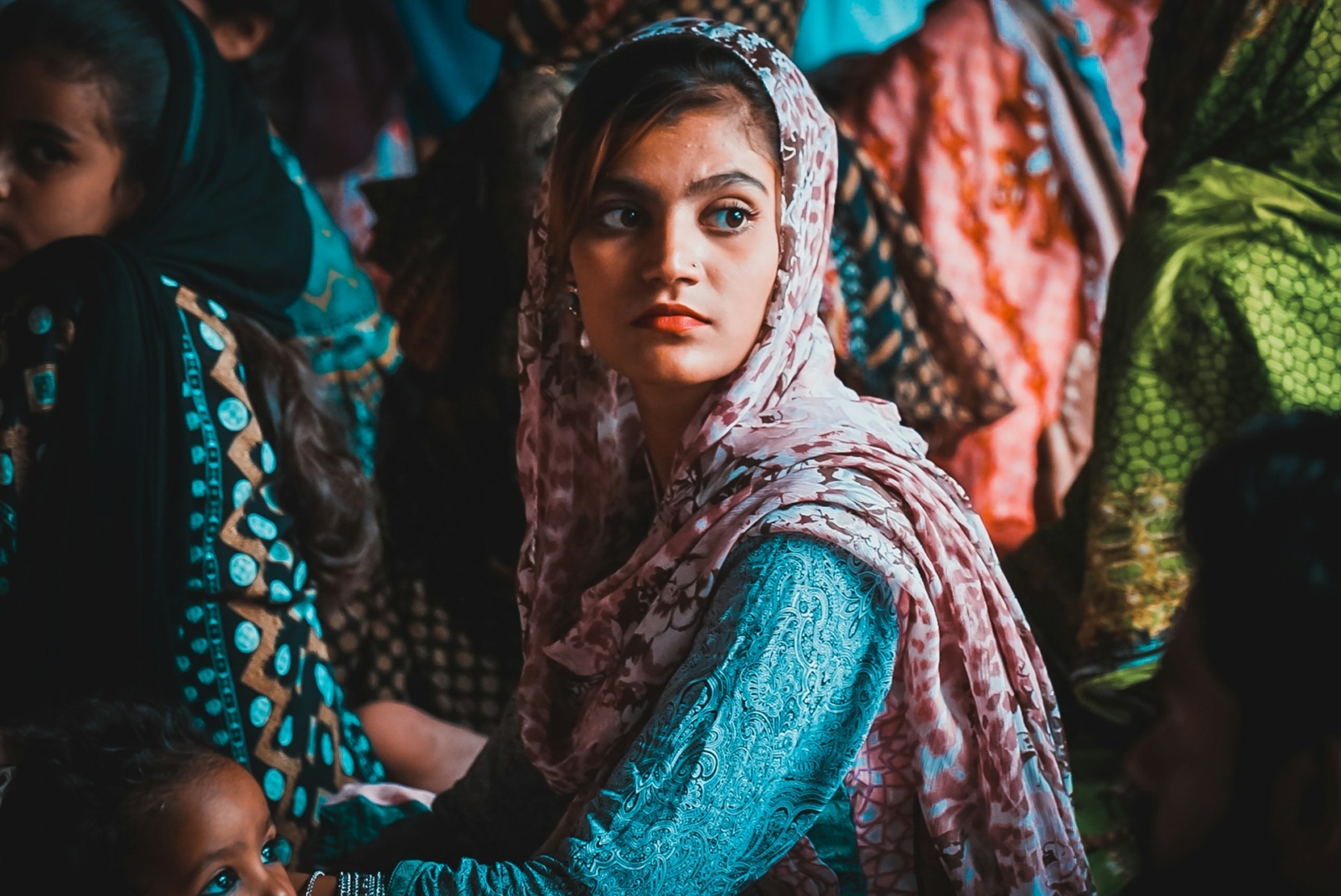 Pakistani woman in a pink head scarf and turquoise dress, sitting on the floor surrounded by other women in brightly coloured clothes.