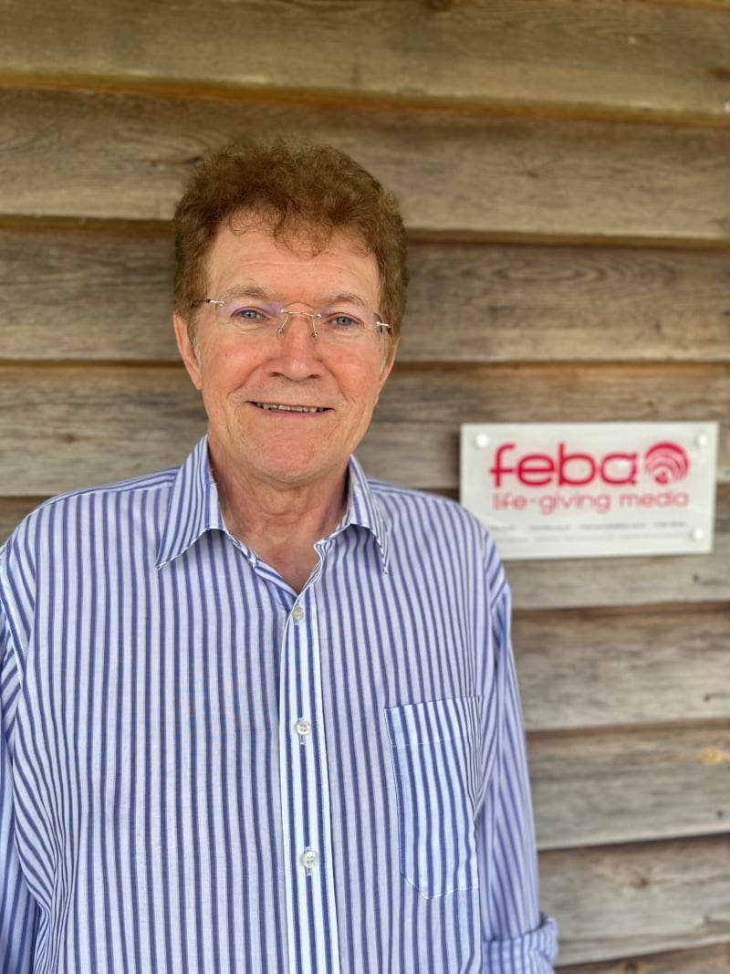 Patrick standing outside the Feba office smiling. He is wearing clear rimmed glasses and a white and blue striped shirt.