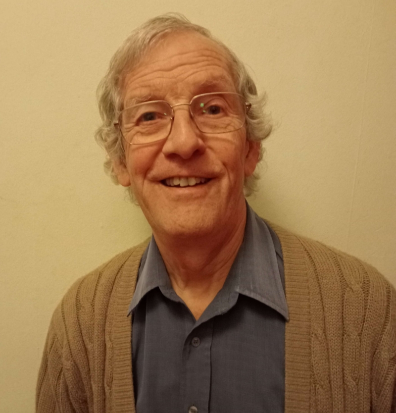 Peter, a man with grey hair and glasses, smiles at the camera. He is wearing a grey shirt with a brown cardigan.