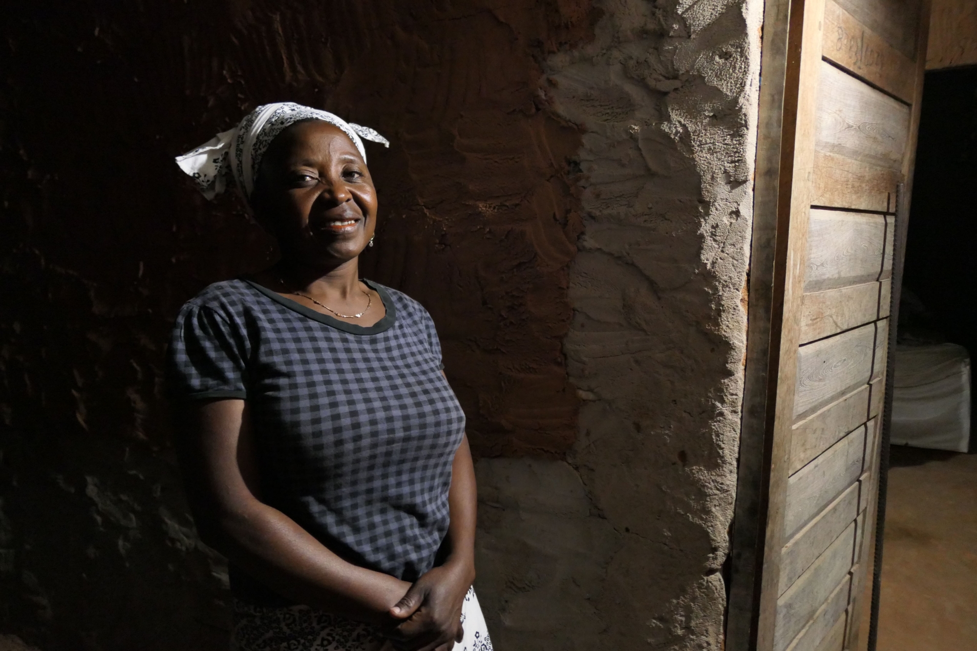 A woman stands in the entrance of her home in Mozambique smiling. She is wearing a white scarf tied on her head and a gingham navy and black tshirt.