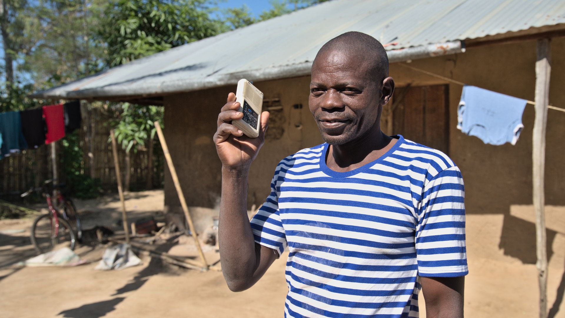 A smiling man standing outside a rural house in Mozambique holding a portable audio device up to his ear.