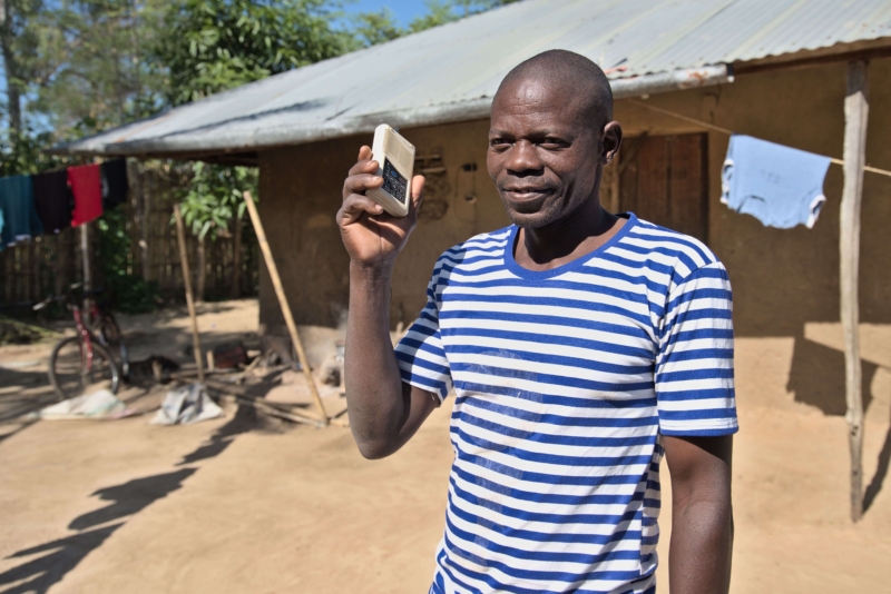 A smiling man standing outside a rural house in Mozambique holding a portable audio device up to his ear.