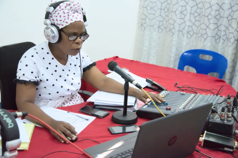 A Radio Wimbe staff member recording a radio broadcast sits at a table covered in a red table cloth in front of a microphone, laptop and sound desk. She is wearing headphones and matching white and black polka dot top and head wrap.