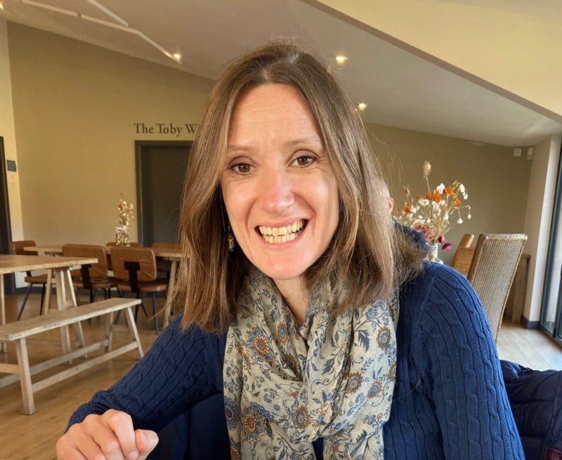 Ruth sitting in a cafe smiling. She had shoulder length brown hair and is wearing a navy jumper with flower patterned scarf.