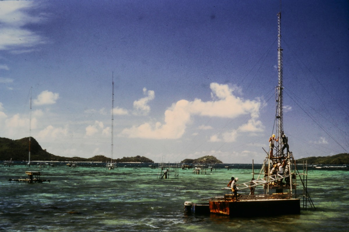 An old film photograph of a radio mast under construction in the ocean in the Seychelles on a sunny day.