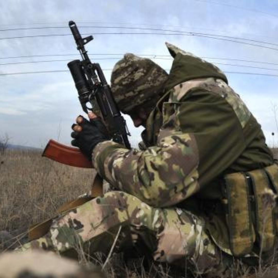 A Ukrainian soldier sitting in a field in full combat gear. He is holding his gun, bent over in prayer.