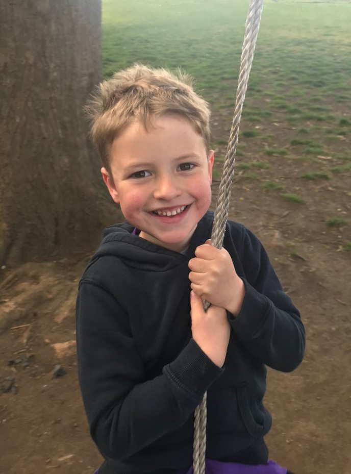 William, a young boy with sandy brown hair, smiling. He is holding a rope swing with a tree in the background.