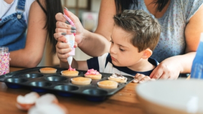 A young boy piping pink icing onto vanilla cupcakes in a baking tray. A woman stand behind him, helping hold the icing bag.