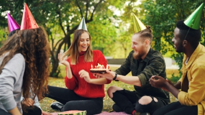 A young woman in a party hat is celebrating her birthday with friends in park on a picnic blanket. A friend holds out a cake with candles as they all smile and laugh.