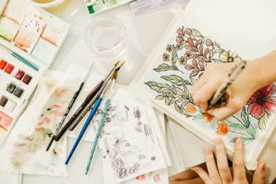 Overhead shot of hands drawing flowers in a sketchbook on a desk surrounded by paintbrushes, scrap paper and watercolour paints.