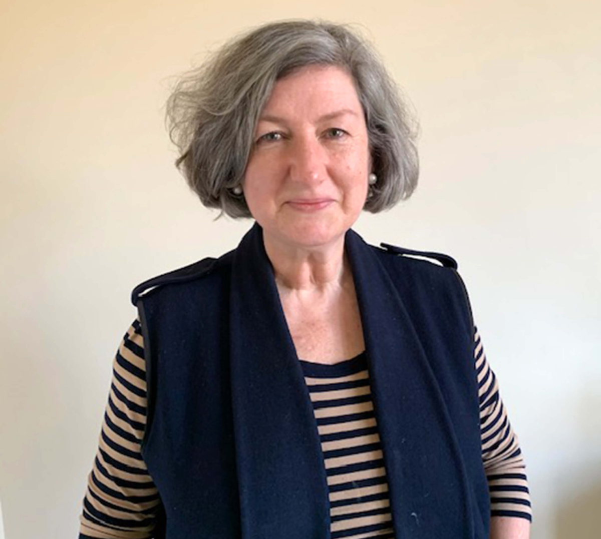 Jo stands against a beige wall smiling. She has a short grey bob, striped tshirt and navy vest with her hands in her pockets.