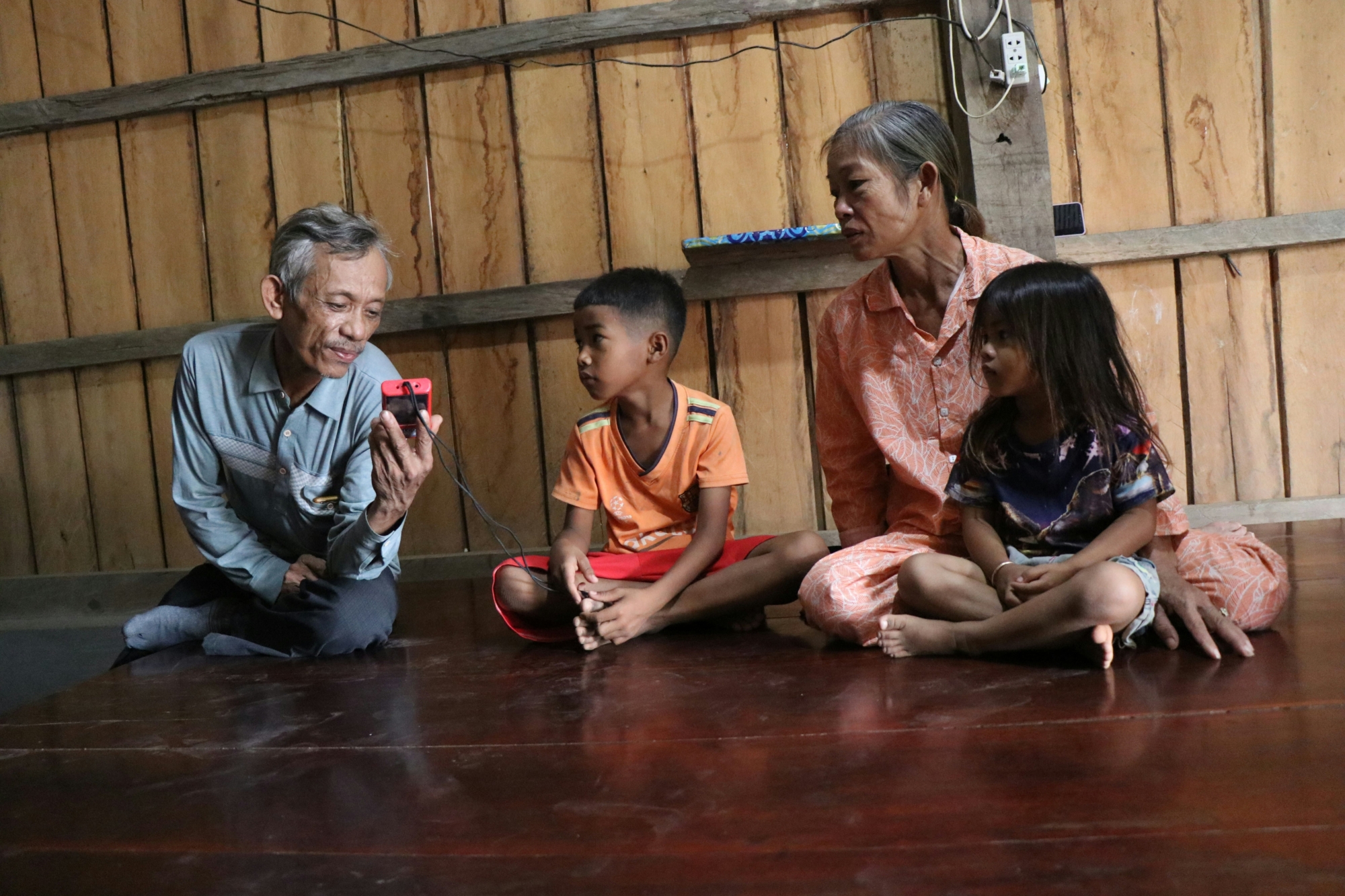 A family in Cambodia sit on on the floor of their wooden house listening to a small red audio Bible.