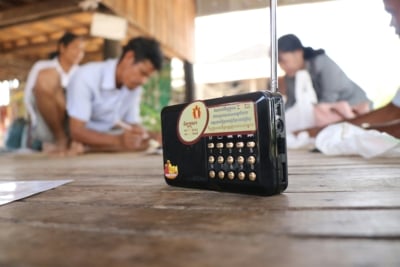 A black radio sits on a wooden floor of a traditional house in Cambodia. In the background are blurred figures sitting and listening.