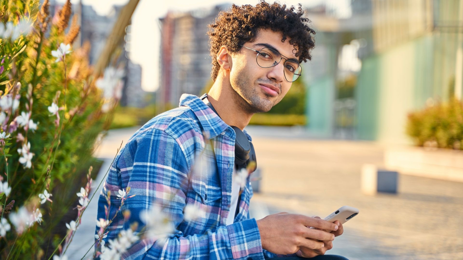 A young man with olive skin and dark curly hair sitting next to a hedge, smiling and holding a phone.