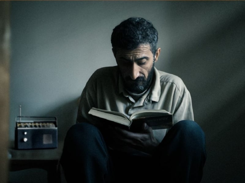 Realistic AI image of an Iraqi man reading a Bible in a dark room, with a radio on a small table beside him.