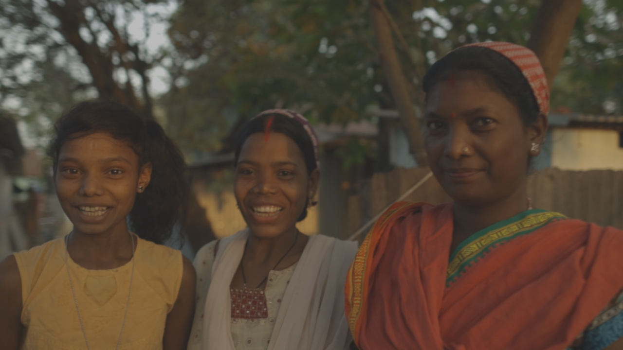 Three young Indian women smile at the camera wearing colourful clothes. There are trees and simple houses in the background.
