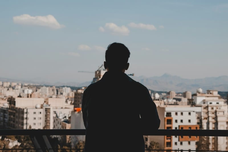 A man standing on a balcony overlooking a city. He is silhouetted against a blue sky with white high rise buildings in the distance.