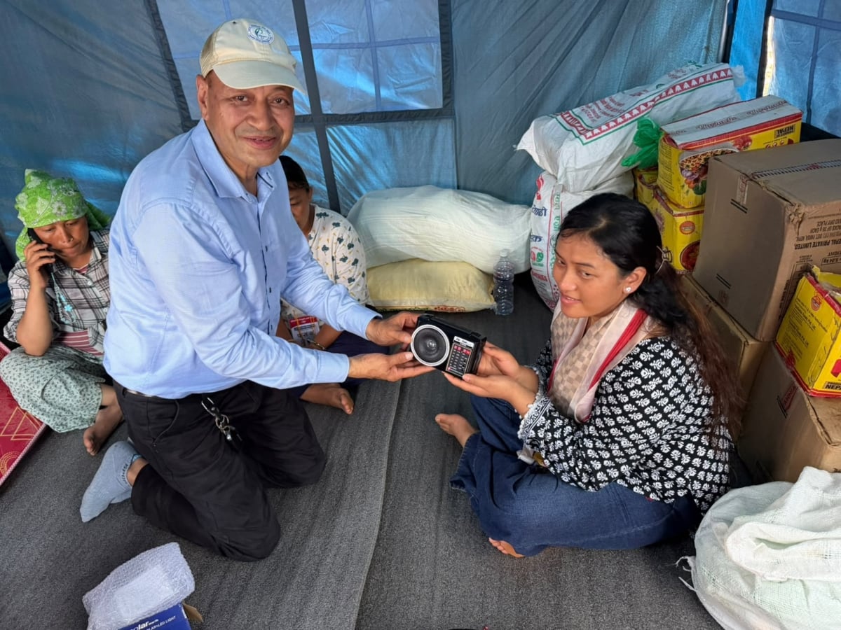 Vijay, director of Afno FM, handing a radio to a young woman affected by the Nepal flood. They are sitting in a temporary shelter surrounded by boxes of food.