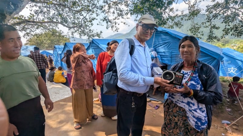 Vijay from Afno FM handing a woman displaced by the 2026 Nepal floods a radio. They are standing together outside with temporary tent shelters in the background and people milling around.