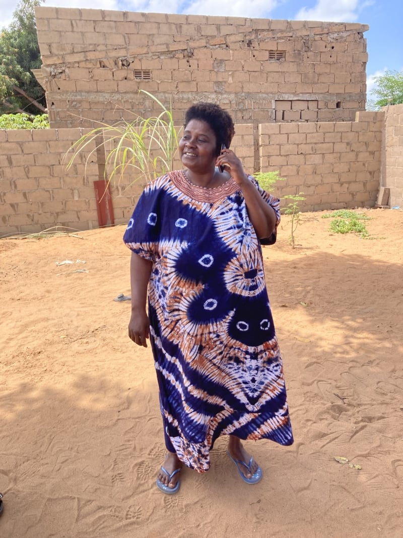 A woman stands in a sandy courtyard in Mozambique holding her phone to her ear, listening to Radio Wimbe's broadcasts. She is wearing a blue and orange tie dye dress and has a short afro.