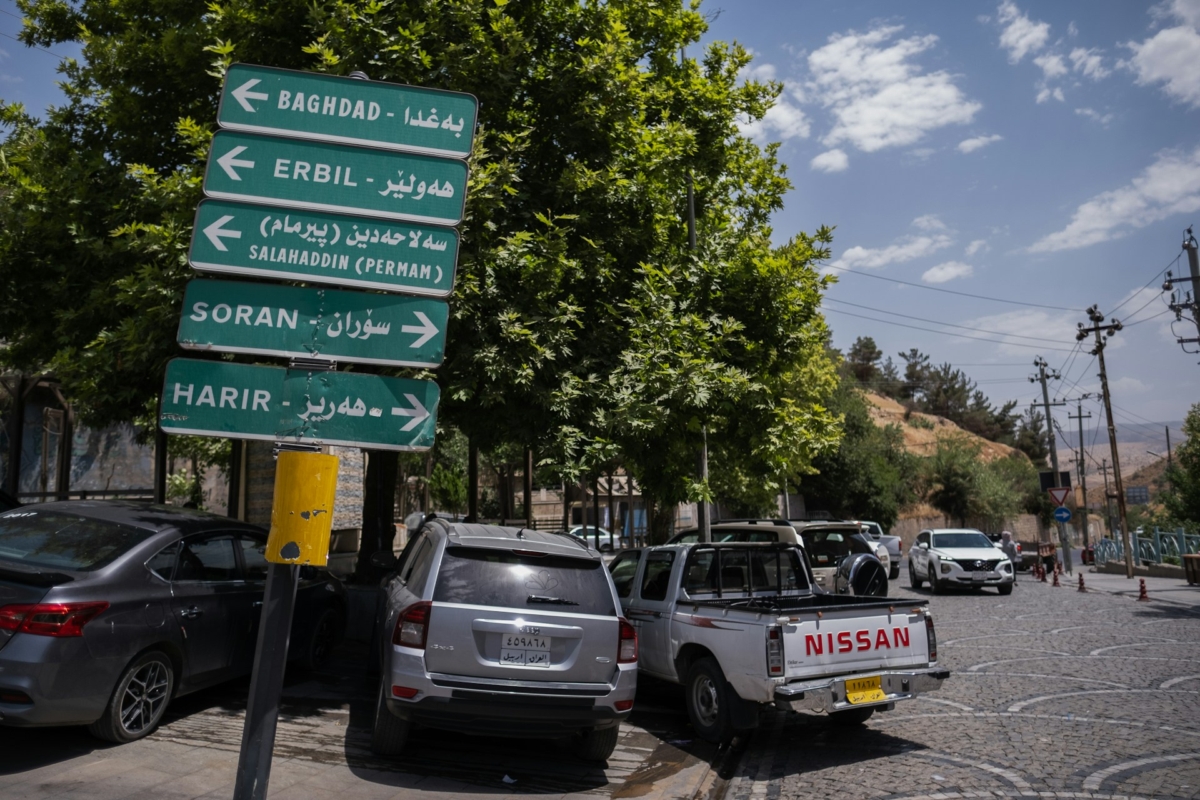 A sunny cobbled and tree lined street in Iraq with cars parked along the road. A green road sign in the foreground shows directions for Baghdad, Erbil, Perman, Soran and Harir in English and Arabic.
