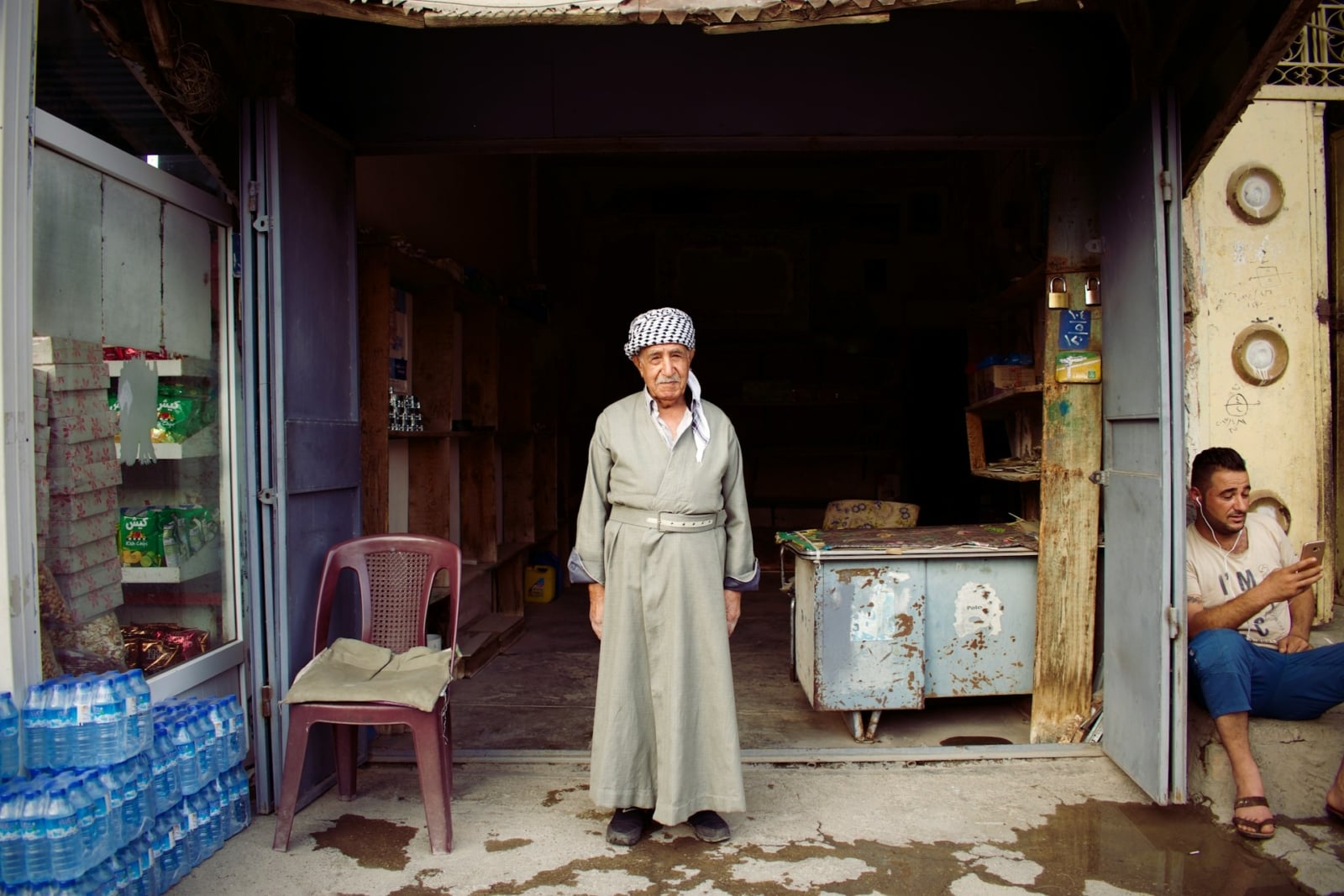 An older man wearing a turban stands outside a simple shop on a street in Iraq.